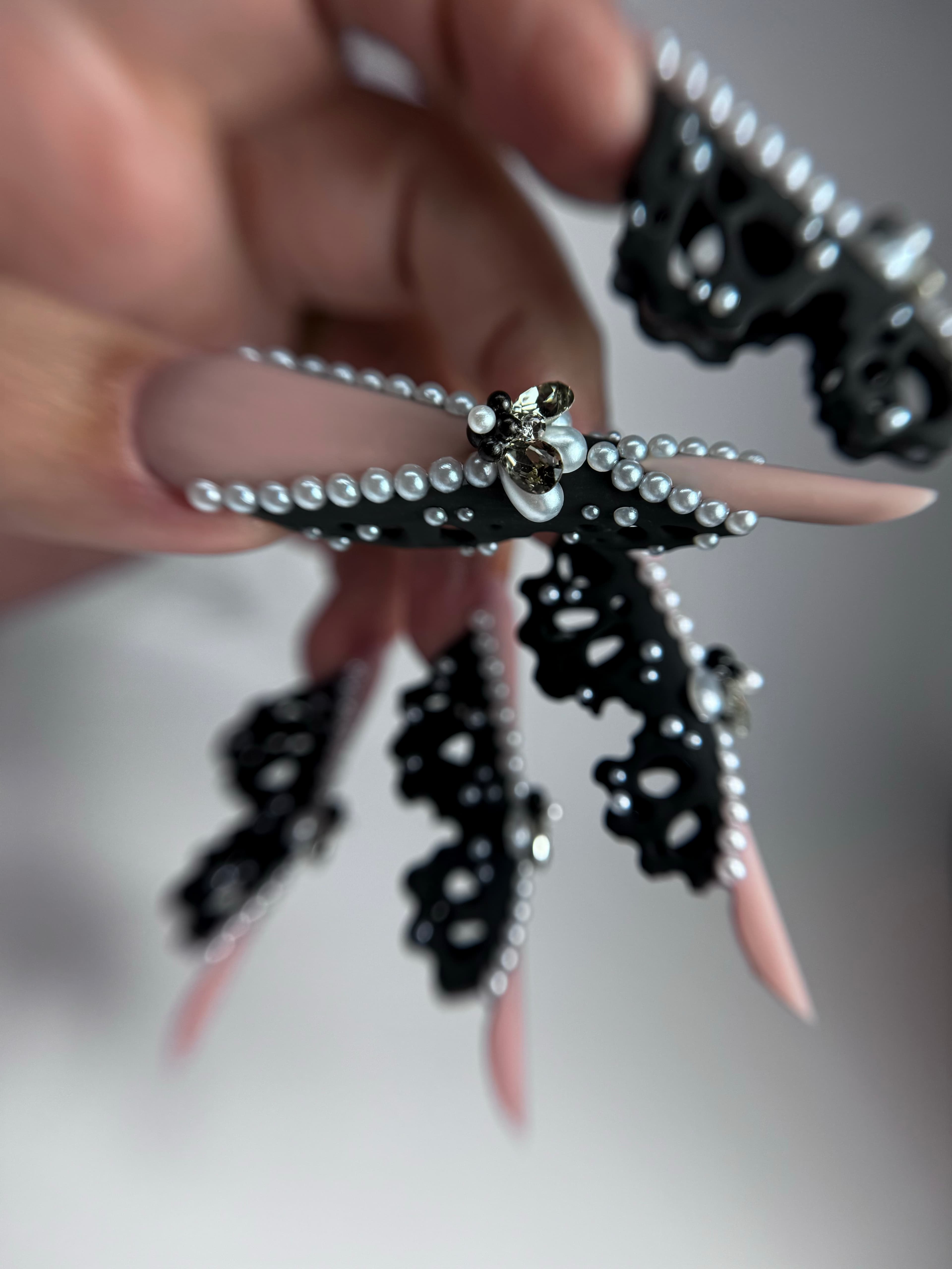 Ongles stiletto nude avec dentelle noire 3D, perles blanches et un bijou central étincelant.