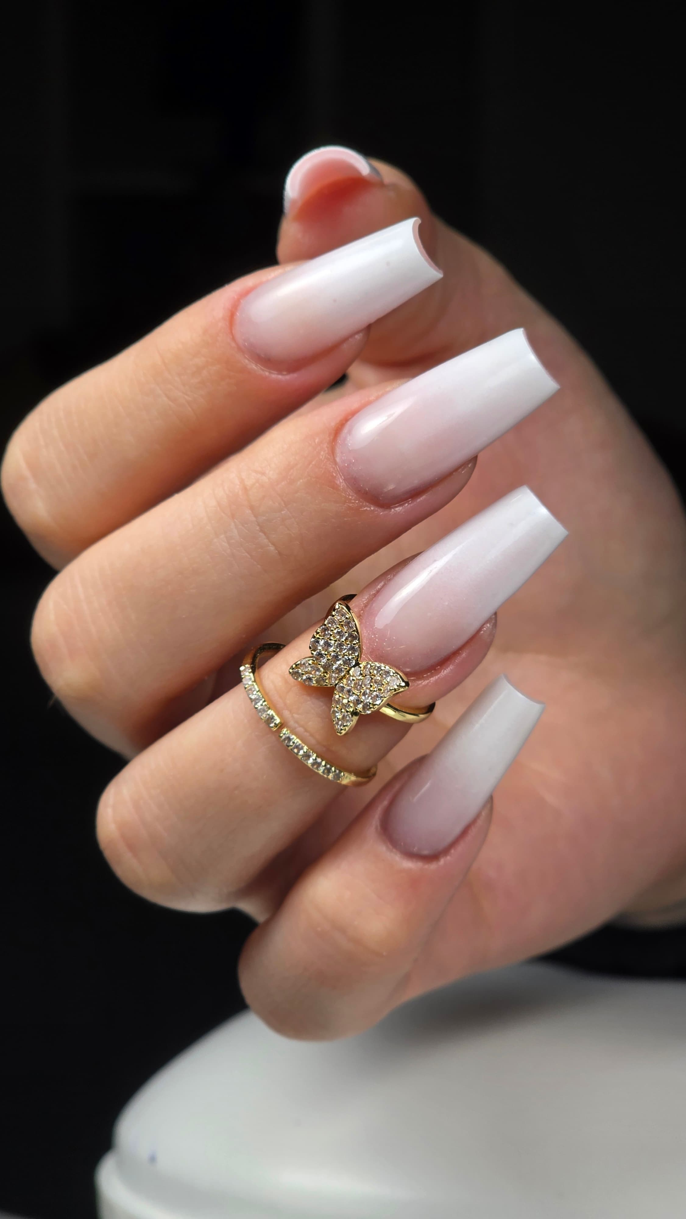 Main aux longs ongles carrés dégradés blancs portant une bague papillon dorée et scintillante.