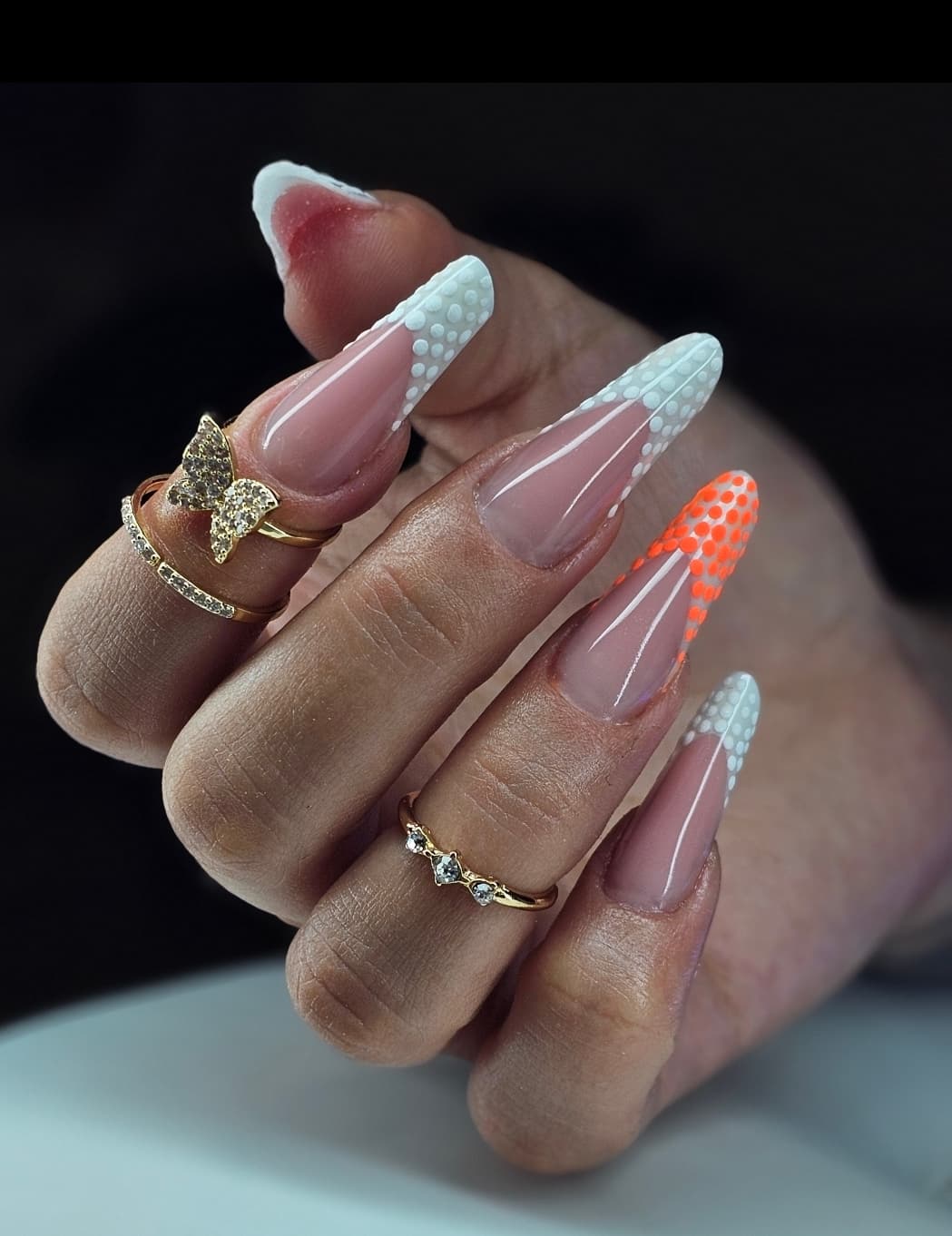 Longs ongles stiletto avec pointes à pois blancs et orange, agrémentés de bagues dorées.