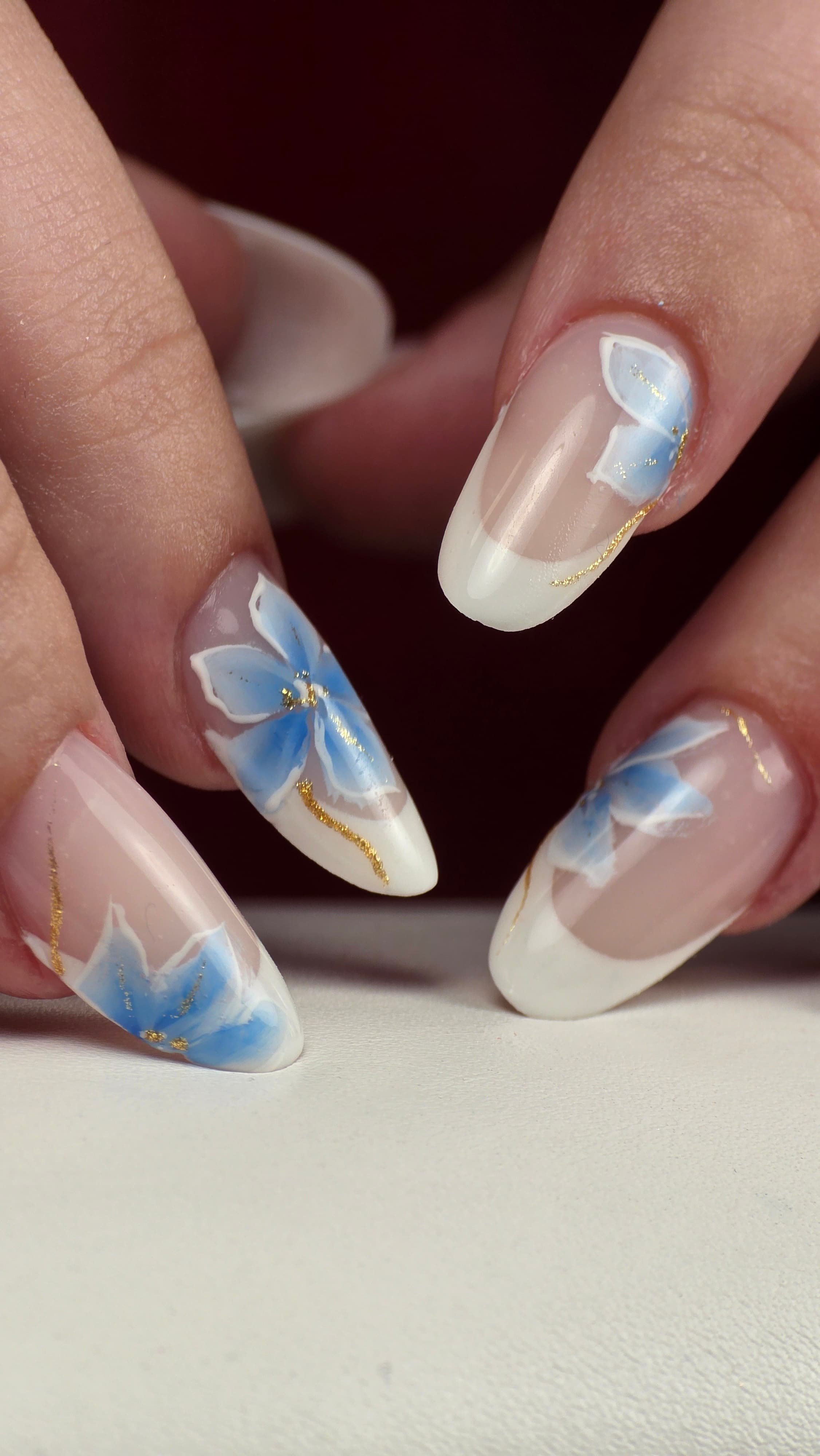 Manucure française sur ongles longs en amande avec motifs de fleurs bleues et accents dorés.