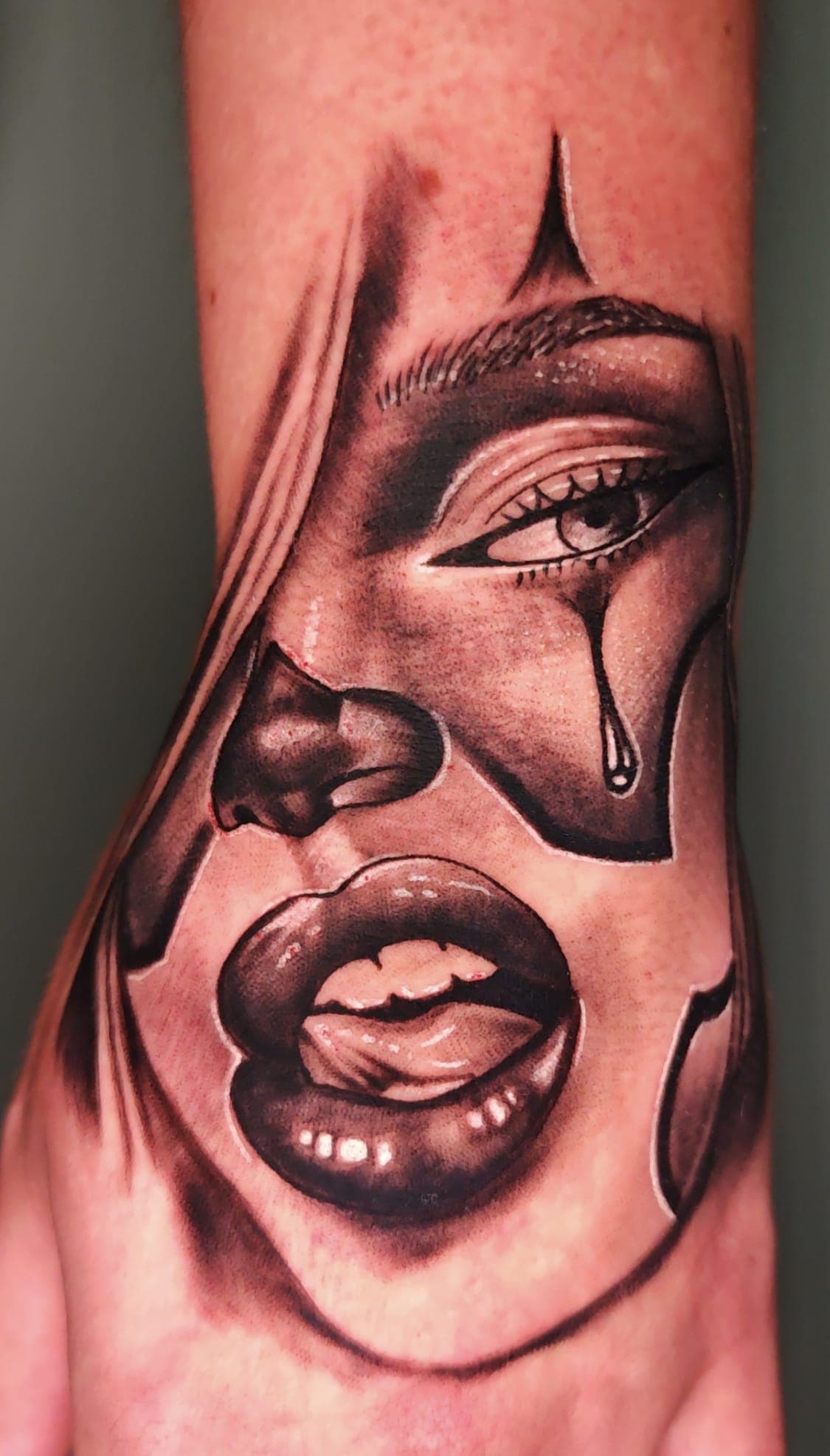 Tatouage noir et gris d'un visage de femme avec maquillage de clown et larme.