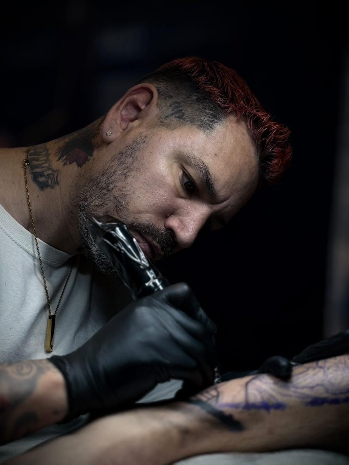 Tatoueur aux cheveux roux et gants noirs travaillant avec précision sur un tatouage complexe.