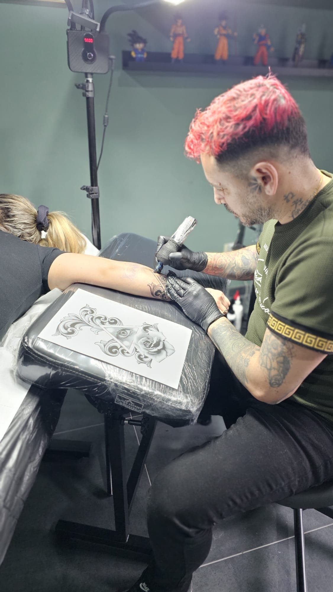 Tatoueur aux cheveux roses tatouant une rose et un œil sur l'avant-bras d'une cliente.