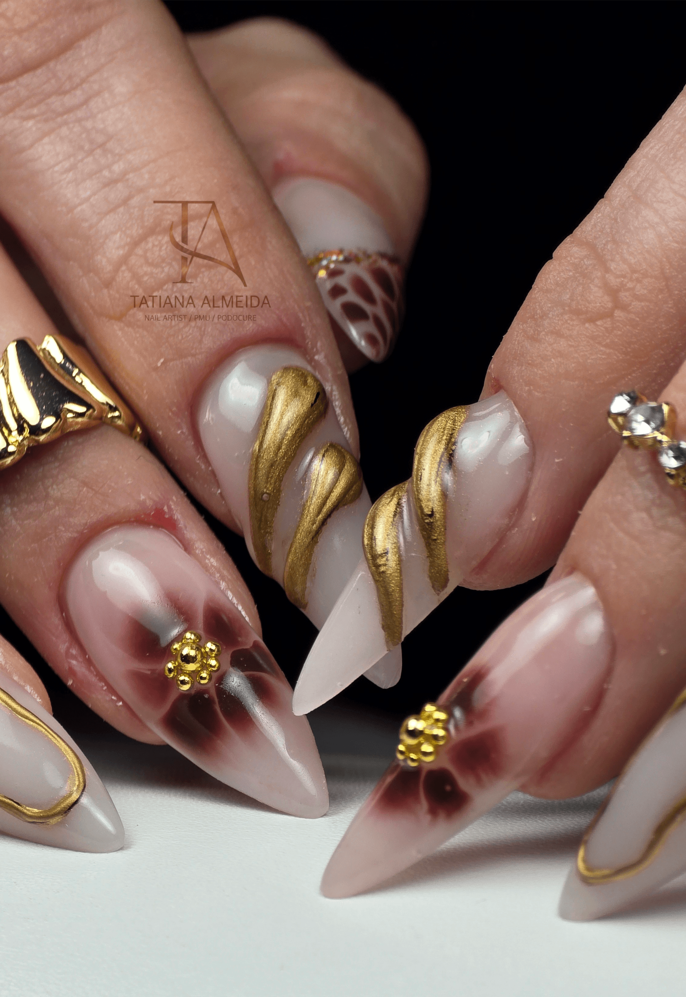 Manucure stiletto avec spirales dorées 3D et fleurs bordeaux sur une base blanc laiteux.